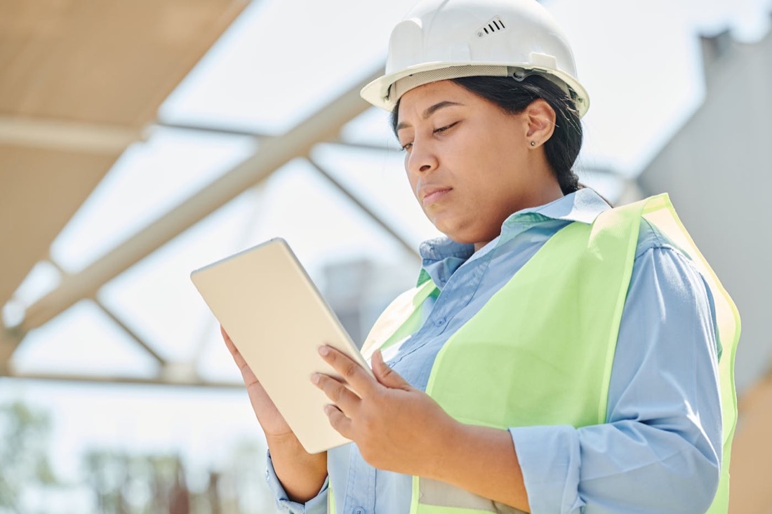 Person mit Schutzhelm und Tablet prueft technische Informationen vor Ort.